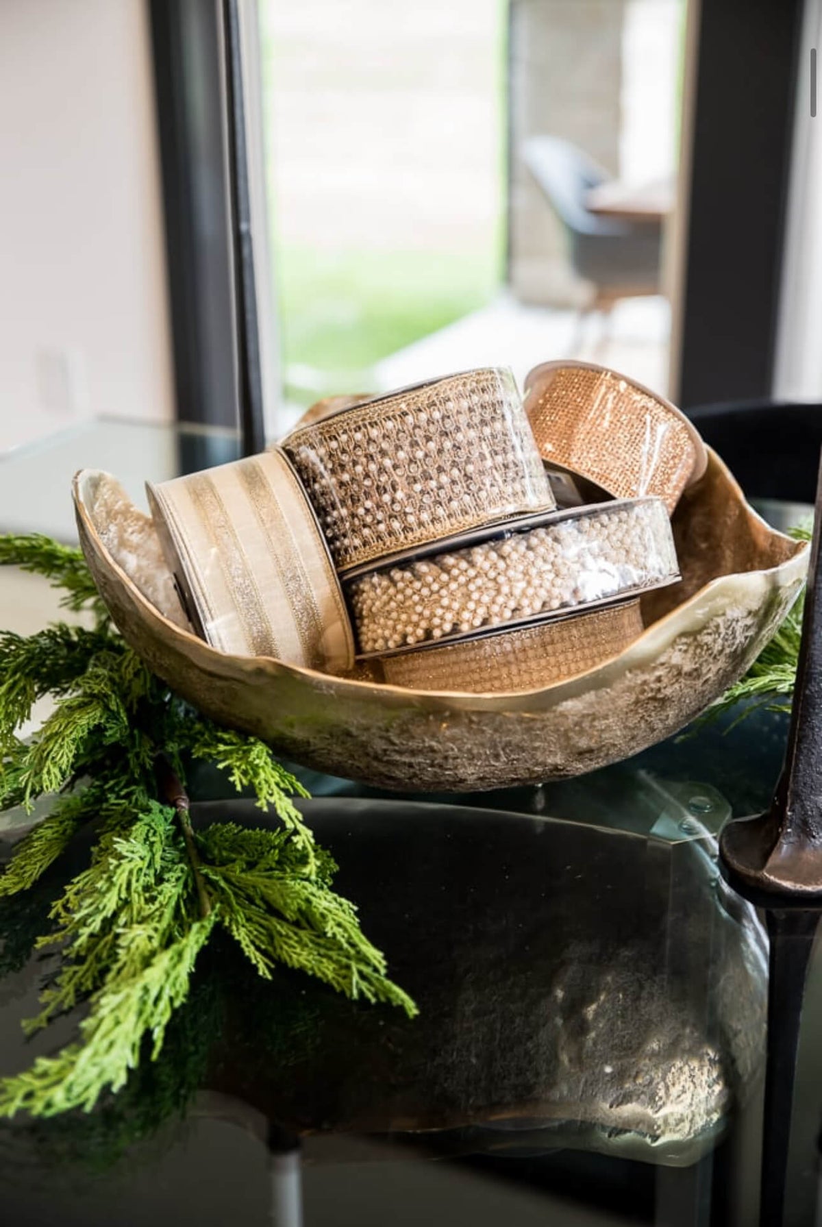 A decorative shell-shaped bowl filled with textured beige and brown decorative ribbons, styled with green foliage on a reflective dark tabletop in an indoor setting.