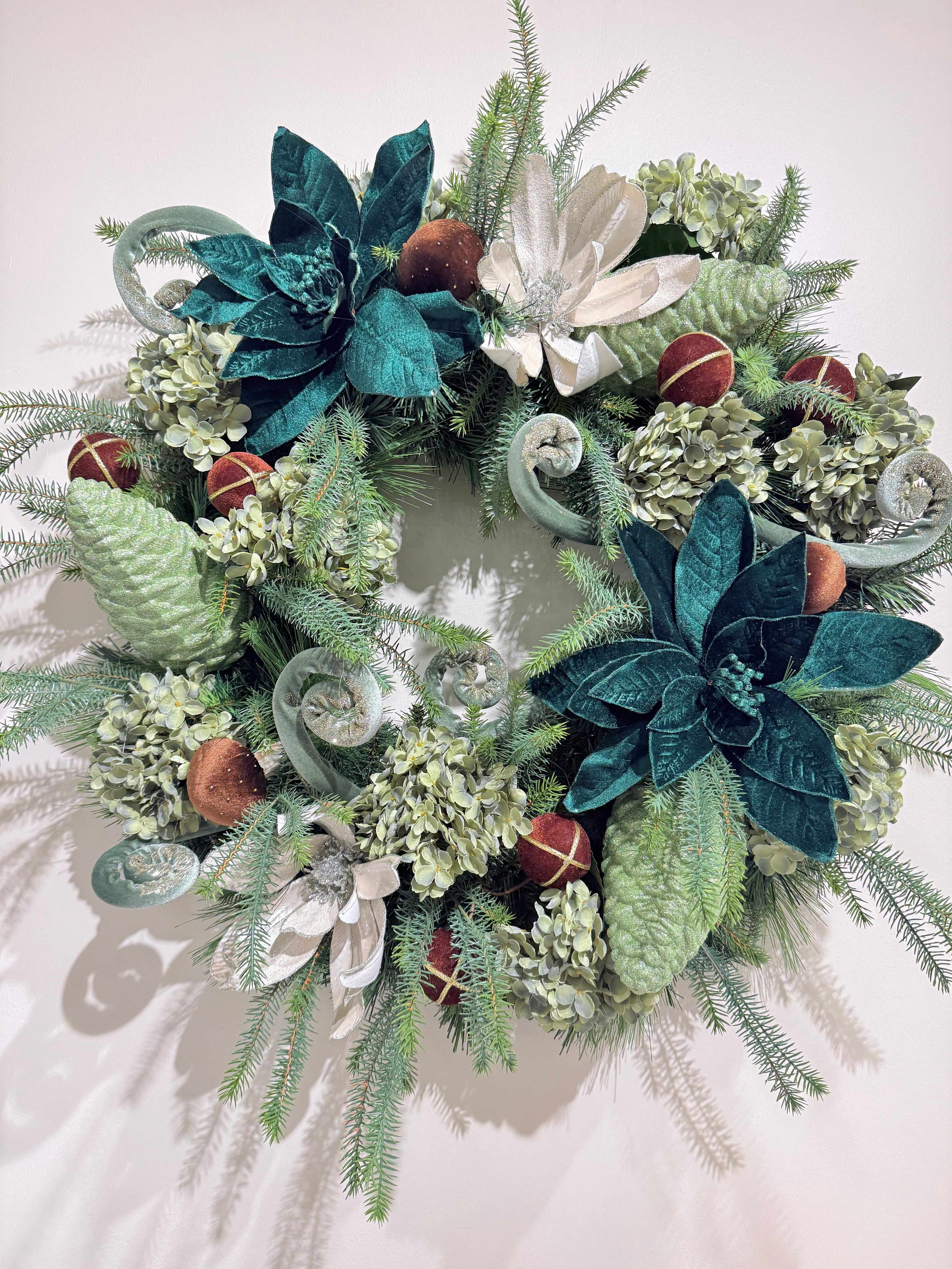 A festive holiday wreath decorated with green pine branches, teal and white artificial flowers, light green pinecones, and small red ornaments, photographed against a plain off-white background.