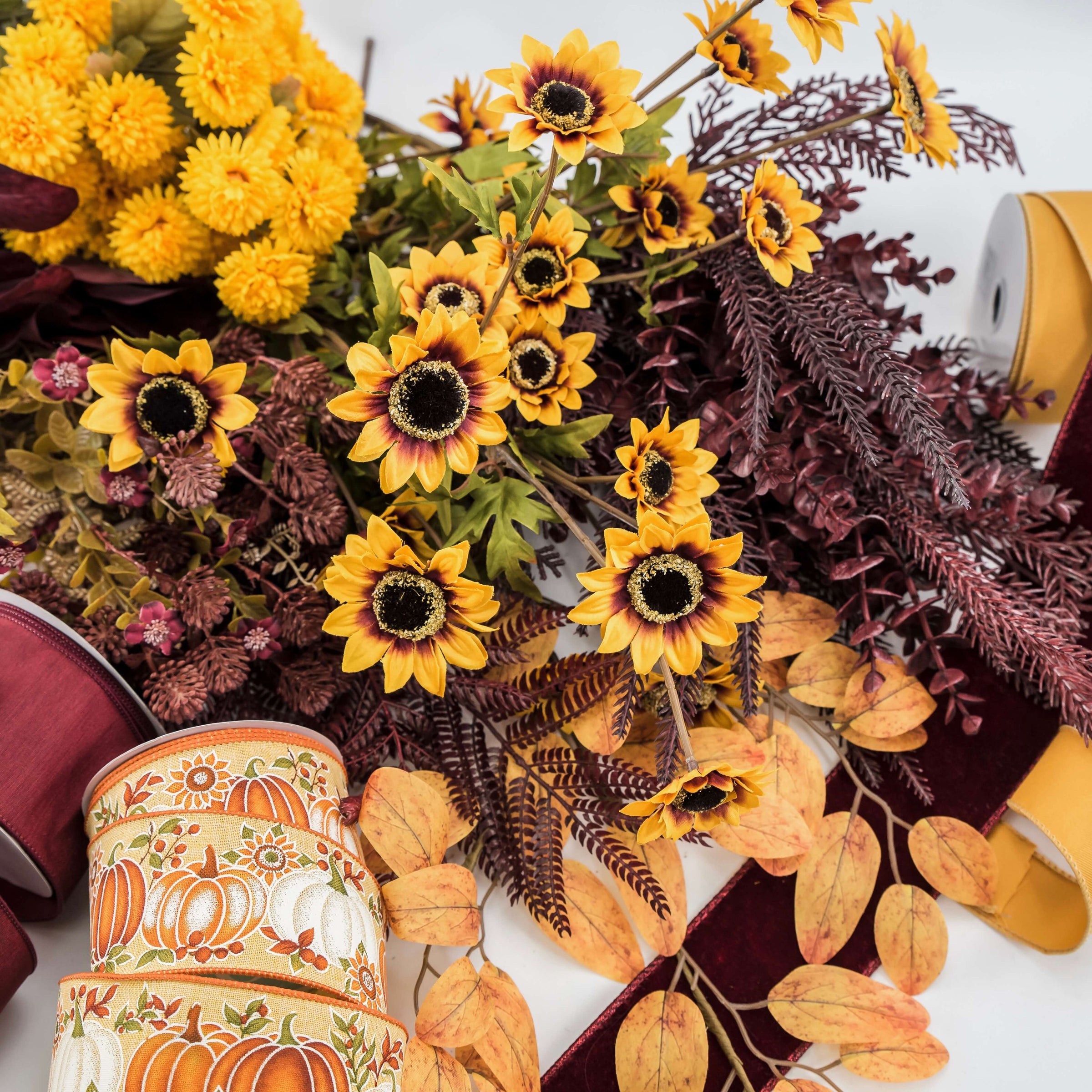 An autumn-themed floral arrangement featuring yellow sunflowers, burgundy and gold ribbons, and fall foliage on a white background.