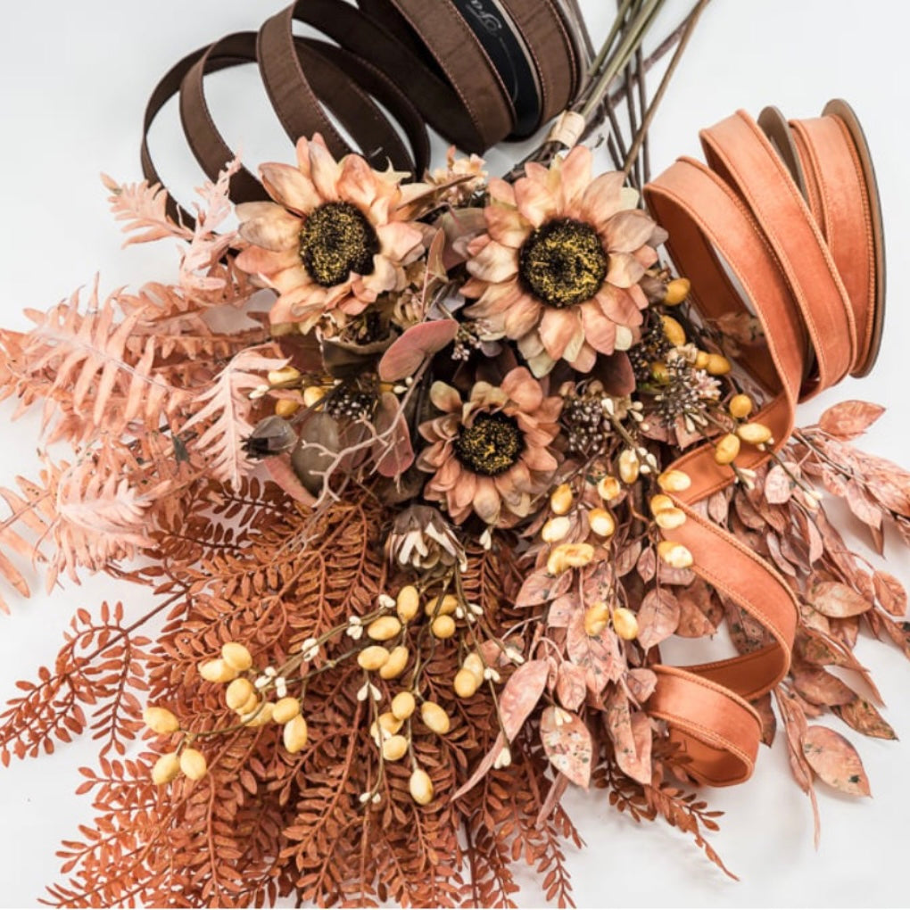 A decorative dried floral arrangement with sunflower-like blooms, rust-colored ferns, small beige berries, and wide curled ribbon spools in brown and terracotta on a white background.