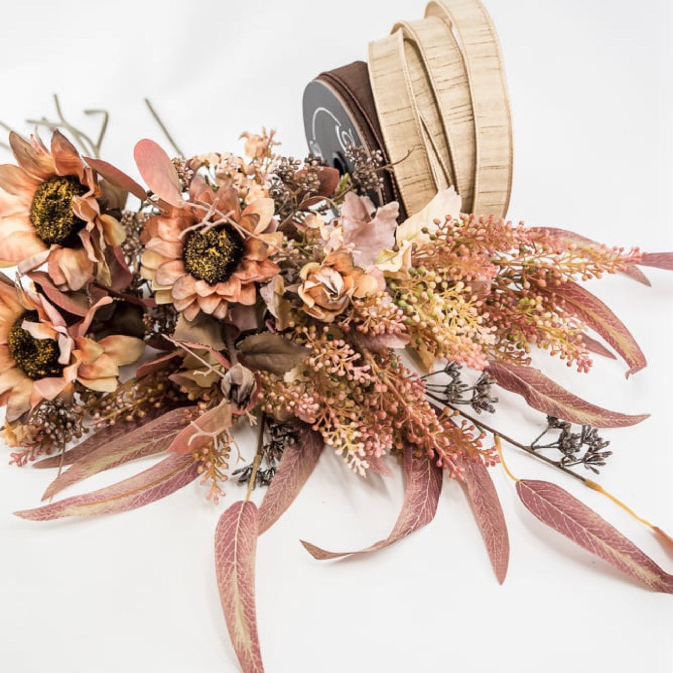 A dried floral arrangement in muted autumn tones with peach, pink, tan, and brown elements, styled with a spool of light wood ribbon on a white background.