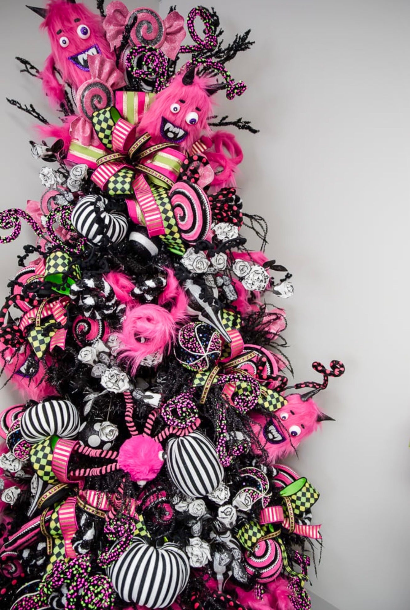 A Halloween-themed decorative tree covered in bright pink and black ribbons, fuzzy monster ornaments, striped pumpkins, black spiderwebs, and spooky ribbons against a plain light gray wall.