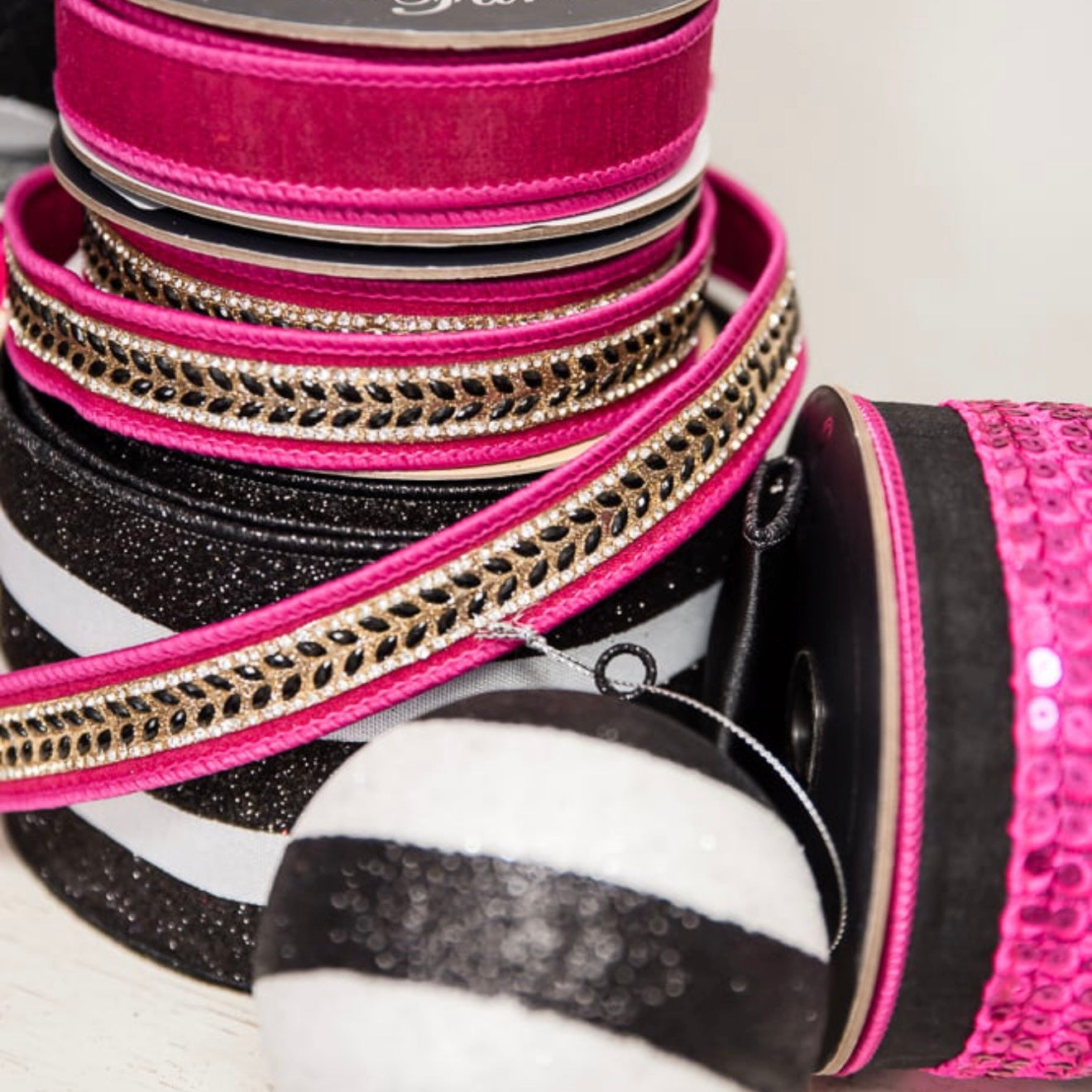 A close-up stack of decorative ribbons and trim spools in bright pink, black, white, and metallic gold, with one spool showing a shiny pink sequined side and another a black-and-white striped ribbon.