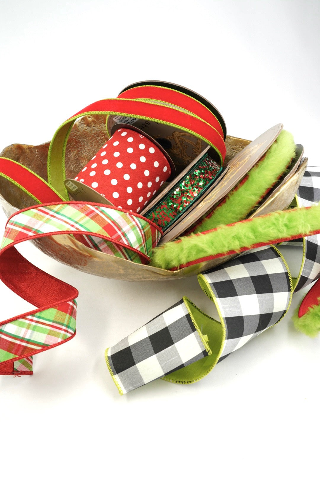 A collection of festive holiday ribbons and decorative trim spools arranged in a shallow wooden bowl on a white background. The ribbons include red with white polka dots, red and green plaid, black and white gingham, and bright green fuzzy trim.