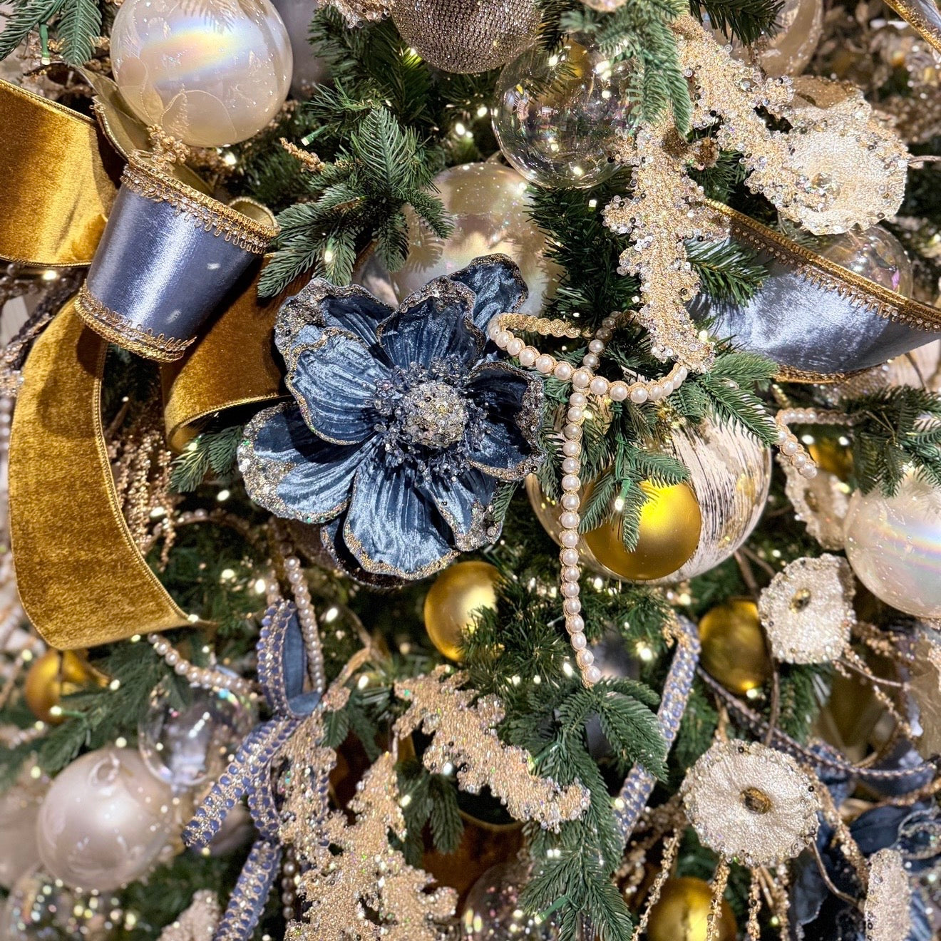 Close-up of a decorated Christmas tree with green pine branches and holiday ornaments, including blue and gold ribbons, pearl-like beads, glittery snowflake ornaments, and iridescent baubles.