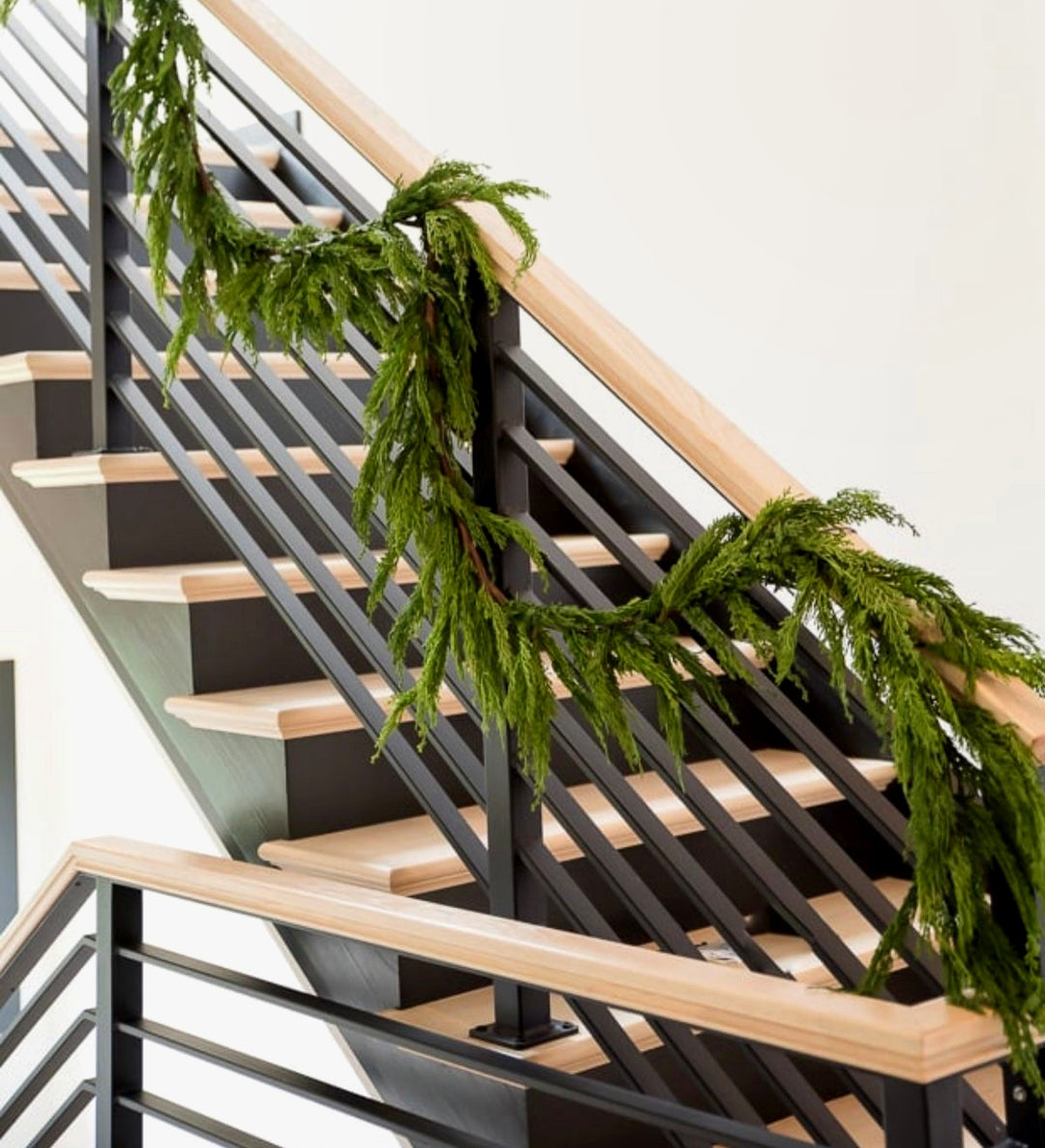 A green evergreen faux garland draped over a modern staircase with black metal railings and light wood steps and handrails.