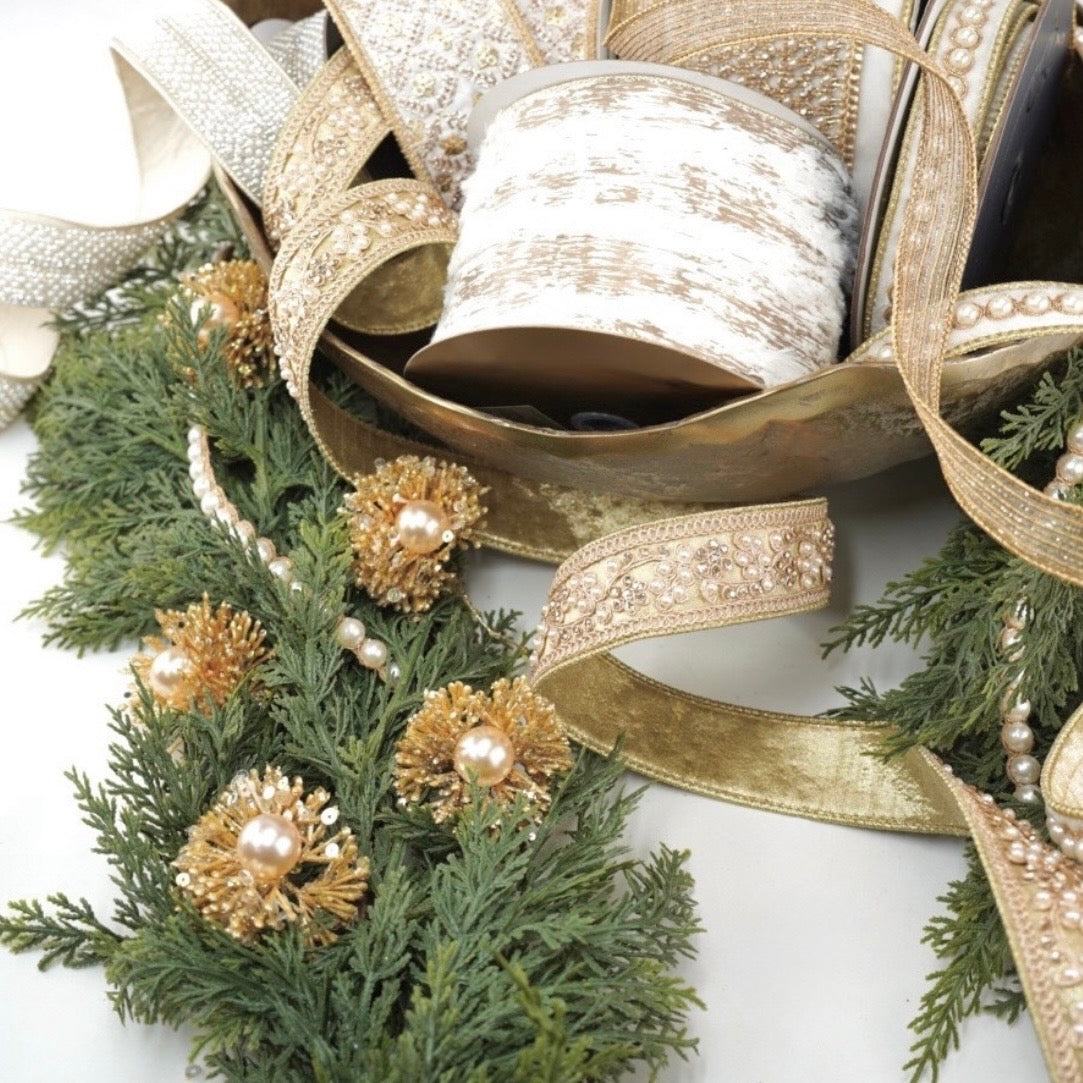 A close-up of a holiday garland and green pine branches, decorated with gold glittered pinecones, pearl beads, and draped gold ribbon with rhinestone trim.
