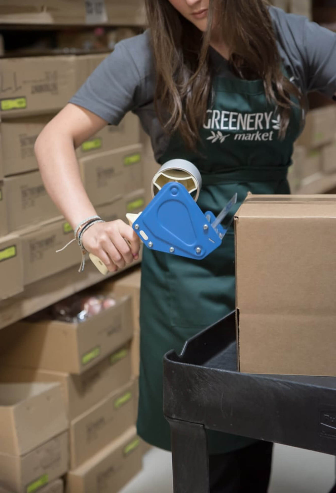 Person in a Greenery Market apron using a box cutter in a warehouse setting
