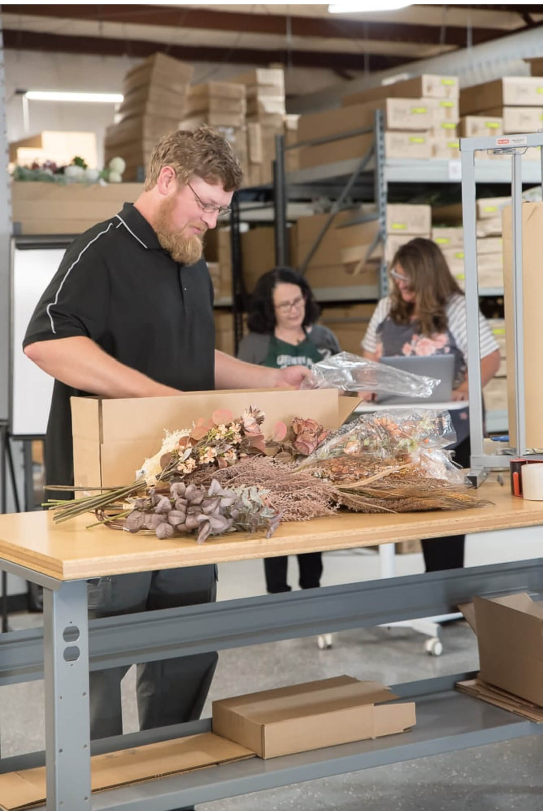 Person packaging flowers in a warehouse setting with colleagues in the background.